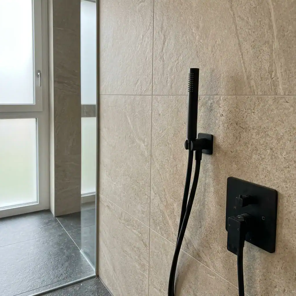 Douchette noire avec flexible fixé sur mur en carrelage beige clair.